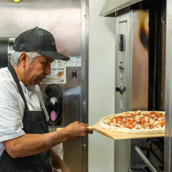 Chef in a black cap and apron sliding a raw pizza with toppings into a commercial oven.