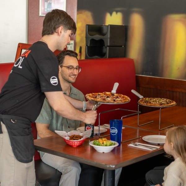 A server places a pizza on a stand for a smiling man and a child at a restaurant booth.