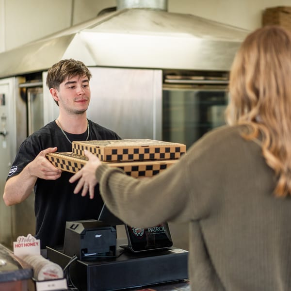 A young man hands two checkered pizza boxes to a customer over a counter.